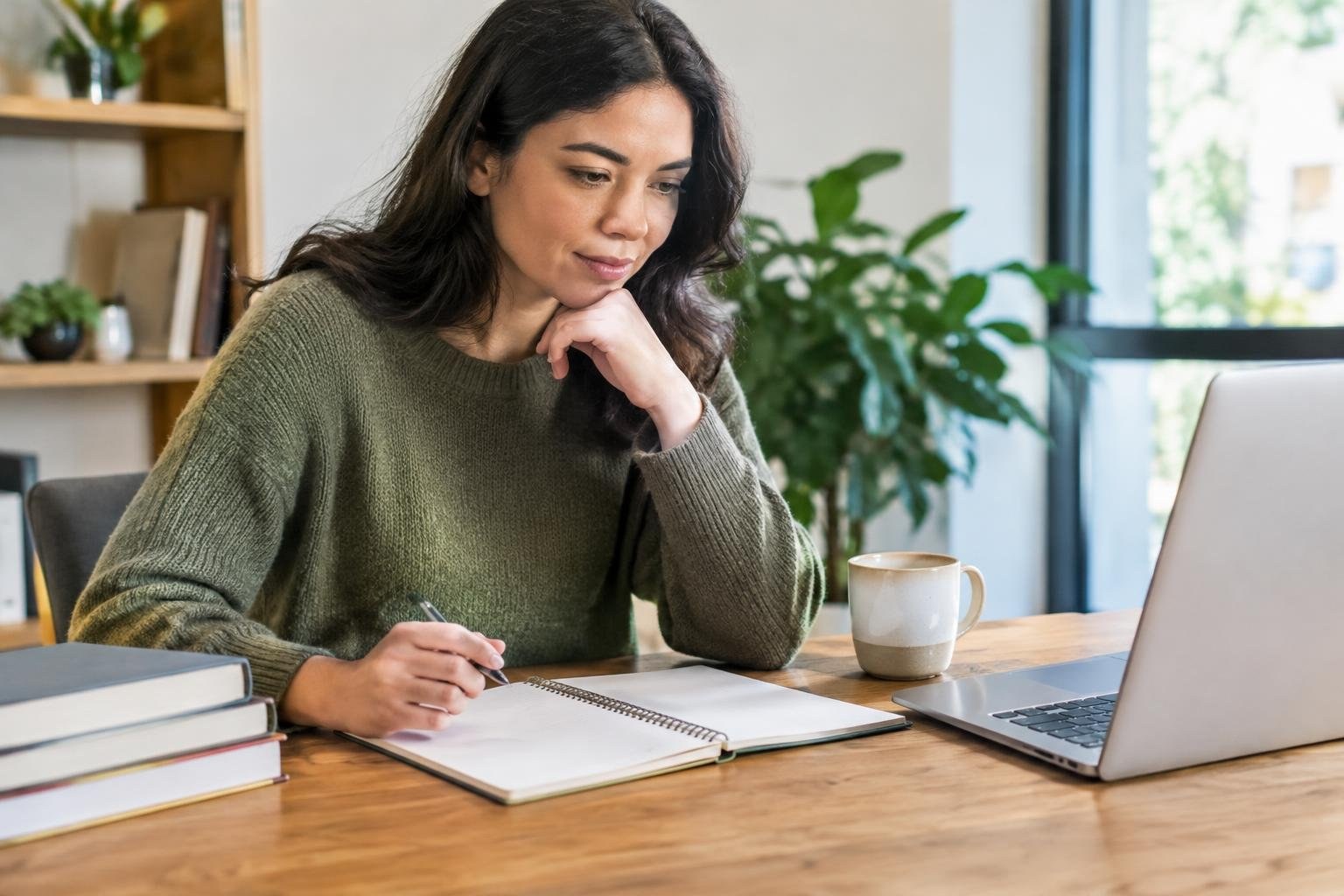 A woman writes in an open notebook at a wooden table beside a laptop, mug and stacked books, with leafy plants and a large window behind her.